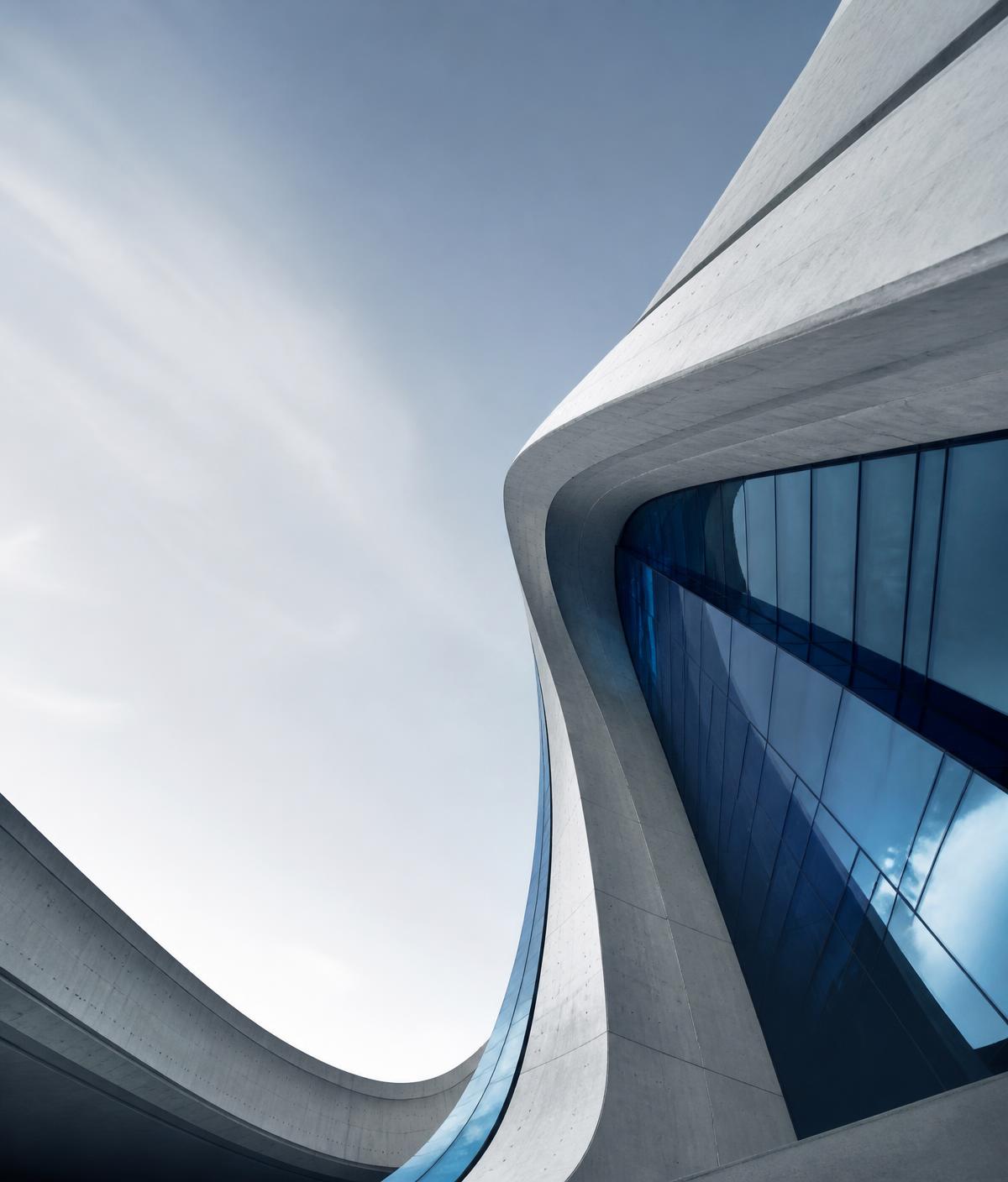Sweeping curves of a large modern concrete and glass building rising into a pale blue sky